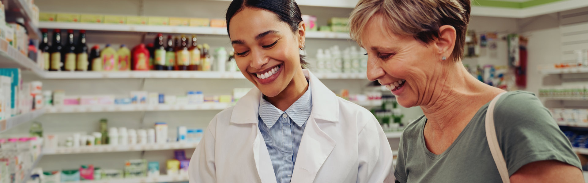 Young female pharmacist and customer reading ingredients and dosage in pharmacy standing near shelves